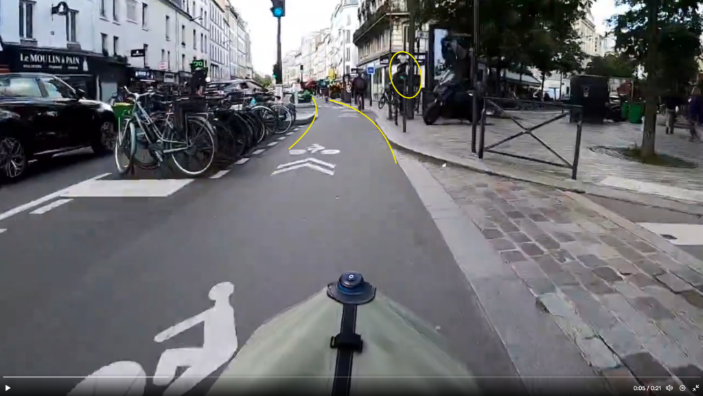 Capture d'écran d'une vidéo de dashcam venant d'un cycliste. Le cycliste est sur une piste cyclable séparée du trafic par un stationnement vélo. À sa gauche, une voie motorisée allant dans le même sens.

Le cycliste a sa voie libre et un feu vert pour le carrefour qui s'annonce.