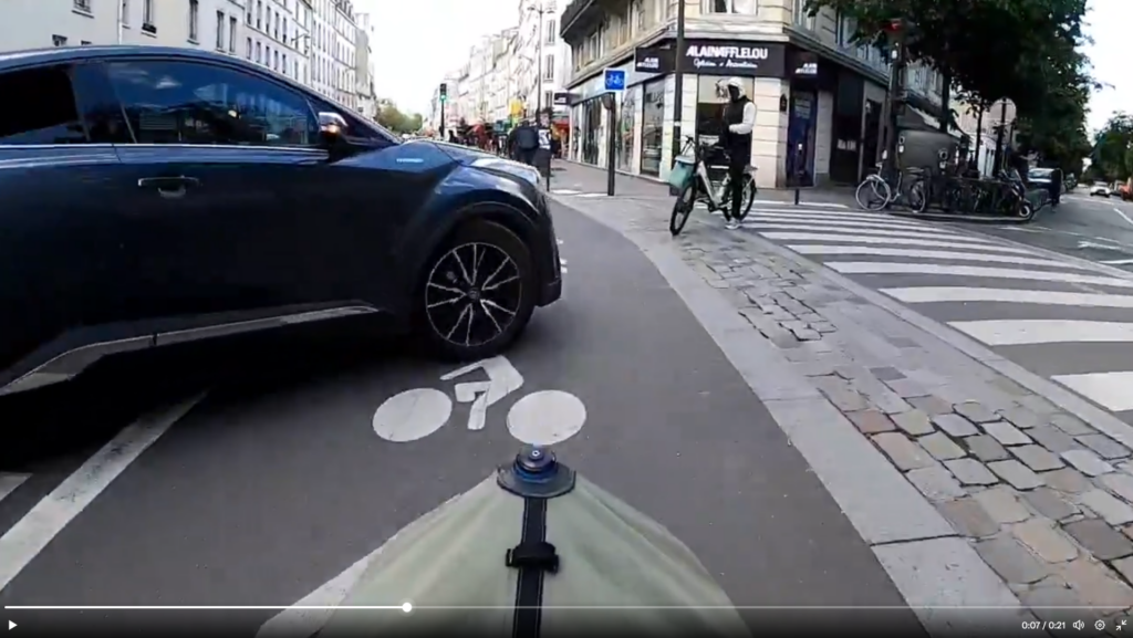 Capture d'écran d'une vidéo de dashcam venant d'un cycliste. Le cycliste est sur une piste cyclable séparée du trafic par un stationnement vélo. 

Au niveau du carrefour, un automobiliste s'est engagé devant lui en venant de la gauche, et bouche le passage à un mètre devant.