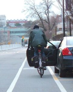 Cycliste roulant sur une bande cyclable. La portière d'une voiture stationnée est ouverte juste devant lui, lui bloquant le passage